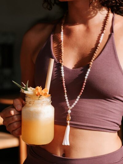 Woman dressed in purple shorts and a soft, comfortable V-neck bra top with delicate straps, drinking a cocktail and wearing a mala necklace.