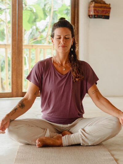 Yogini seated in Sukhasana meditation, wearing comfortable ecru wide-leg pants and a purple yoga top with a subtle V-neck, fitted at the hips.
