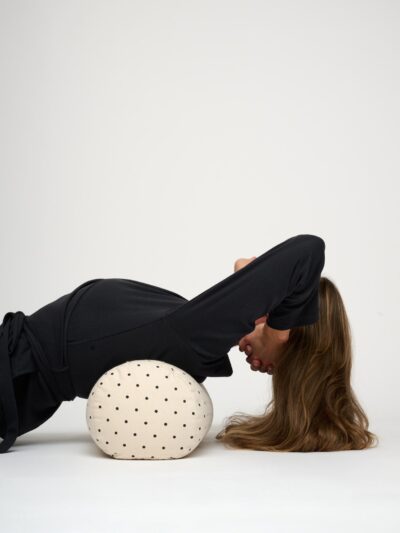 A woman lying on her back, propping her head up with her hands, with a round, ecru yoga bolster with black polka dots placed across her shoulder blades