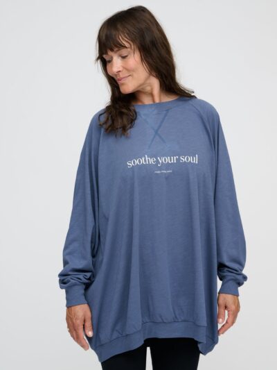 A woman is standing, wearing an indigo blue sweatshirt with white lettering on the front; it is oversized, long, and loose-fitting.